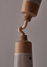 Close-up of a brown tube being squeezed, with beige cream oozing out against a gray background.