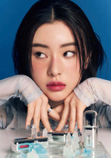 Woman with dark hair and red lipstick sitting at a table with various items including a phone and small bottles.