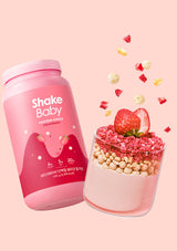 Pink 'Shake Baby' protein shake container with a strawberry-flavored shake in a glass on a pink background