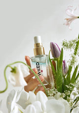 Hand holding a glass bottle with a gold spray nozzle, surrounded by flowers on a light background