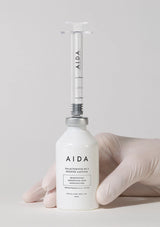 Bottle of AIDA skincare product held by a gloved hand on a light gray background