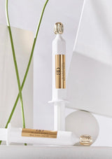 Two skincare products with gold caps on a white surface with green leaves in the background