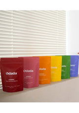 Five Odada product packages in red, pink, orange, yellow, and blue on a white surface with a window blind background.
