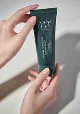 Hand holding a green skincare tube labeled 'mi' against a neutral background