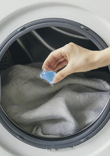 Hand placing a blue laundry detergent pod into an open washing machine.