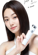 A woman with long, straight dark brown hair holds a white handheld beauty device with a silver and black cap against a clean white background. Korean text and a signature are visible in the upper right corner.