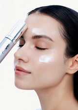 A close-up of a woman with closed eyes, showing a white cream or serum being applied to her forehead with a silver tube, against a clean white background.