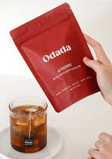 Red Odada tea bag package held by a hand with a glass of iced tea on a white surface.