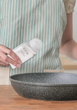 Person holding a bottle of cleaning product next to a frying pan on a wooden surface.