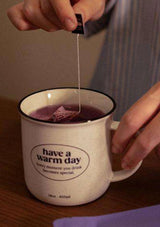Person adding a tea bag to a mug with 'have a warm day' text.