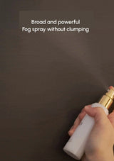 Hand holding a white spray bottle with gold nozzle on a dark background, demonstrating fog spray.