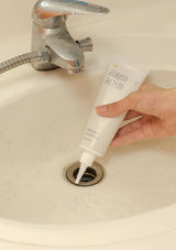 Hand using a tube of white cream to unclog a sink drain.