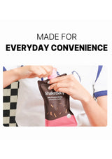 A person holding and opening a Shakebaby milk protein shake pouch in chocolate flavor, with large text above reading 'MADE FOR EVERYDAY CONVENIENCE' on a white background.