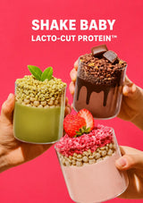 Three hands hold up three different protein shake parfaits against a bright pink background with the text 'SHAKE BABY LACTO-CUT PROTEIN' at the top. The left shake is green with green flakes and a mint leaf, the middle-right shake is dark brown chocolate with chocolate drizzle and a chocolate piece, and the bottom shake is light pink with pink flakes and a strawberry.