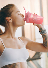 A woman in a white sports bra drinking from a pink ShakeBaby Bottle shaker cup with a straw, set against a softly blurred indoor background.