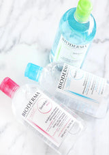 Three Bioderma skincare bottles arranged on a white marble surface: a clear bottle with a pink cap and pink text for Santé-Firming Hand Cleanser, a clear bottle with a blue cap and blue text for Hydroal Hydrating Face Cleanser, and a blue bottle with a green cap for Solisyn Blue Cleanser.