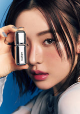 A close-up lifestyle shot of a model holding a silver rectangular cosmetic compact with the brand name 'Odshtye' printed vertically on the front, set against a vibrant blue background.