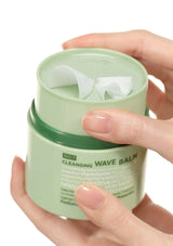 Hand holding a green jar of cleansing wave balm with tissue papers inside.