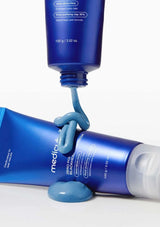 Blue skincare product being squeezed from a tube onto another blue tube with a white cap on a light gray background.