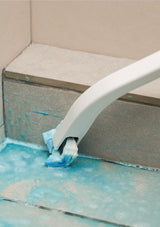 Cleaning tool with blue bristle brush cleaning a tiled wall corner.