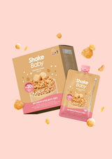 Shake Baby protein powder packaging, showing a cardboard box and a single squeeze pouch with a pink cap, floating on a solid light pink background with scattered cereal pieces.