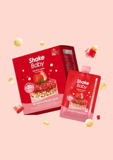 Shake Baby protein powder strawberry flavor shown with a red retail box and a matching red single-serve pouch, floating on a pale pink background with scattered strawberry pieces and cream-colored capsules.