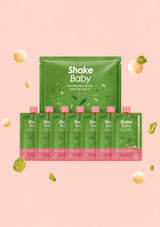 A set of Shake Baby dietary protein shake pouches and bottles arranged on a soft pink background with floating snack pieces. The main product is a large green pouch behind six matching squeeze bottles, all labeled Shake Baby, with green and pink packaging and white text.