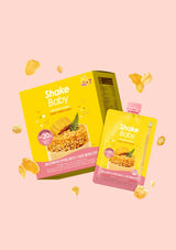 Shake Baby baby food pouch and matching box packaging in a bright yellow and pink color scheme, floating against a solid pale pink background with scattered snack pieces.