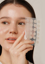 Woman holding a sheet of adhesive patches over her face against a neutral background
