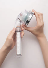 A person's hands holding a transparent cylindrical shower head filter cartridge with white filter blocks inside, while unscrewing a clear and silver shower head attachment against a plain light background.