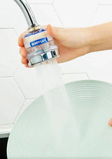 A hand holding a chrome faucet aerator filter with water flowing from it, shown above a plate in a kitchen setting with white hexagonal tile backsplash. The filter has a blue and white label with the brand name 'BODY LUV'.