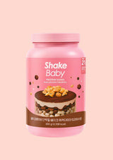A pink container of Shake Baby Protein Shake in Macadamia Tiramisu flavor, featuring a chocolate cake graphic on the label, set against a solid pale pink background.