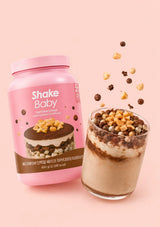A pink container of Shake Baby Protein Shake in Macadamia Tiramisu flavor, featuring a chocolate cake graphic on the label, set against a solid pale pink background.
