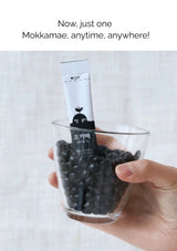 Hand holding a glass of black beans with a Mokkamae product inside, against a light background.