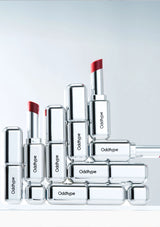 Collection of clear Oddtype lipsticks with red caps on a light gray background