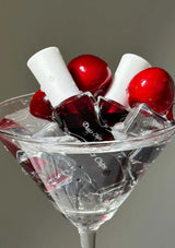 Nail polish bottles with red caps and clear liquid in a martini glass with ice and cherries on a gray background