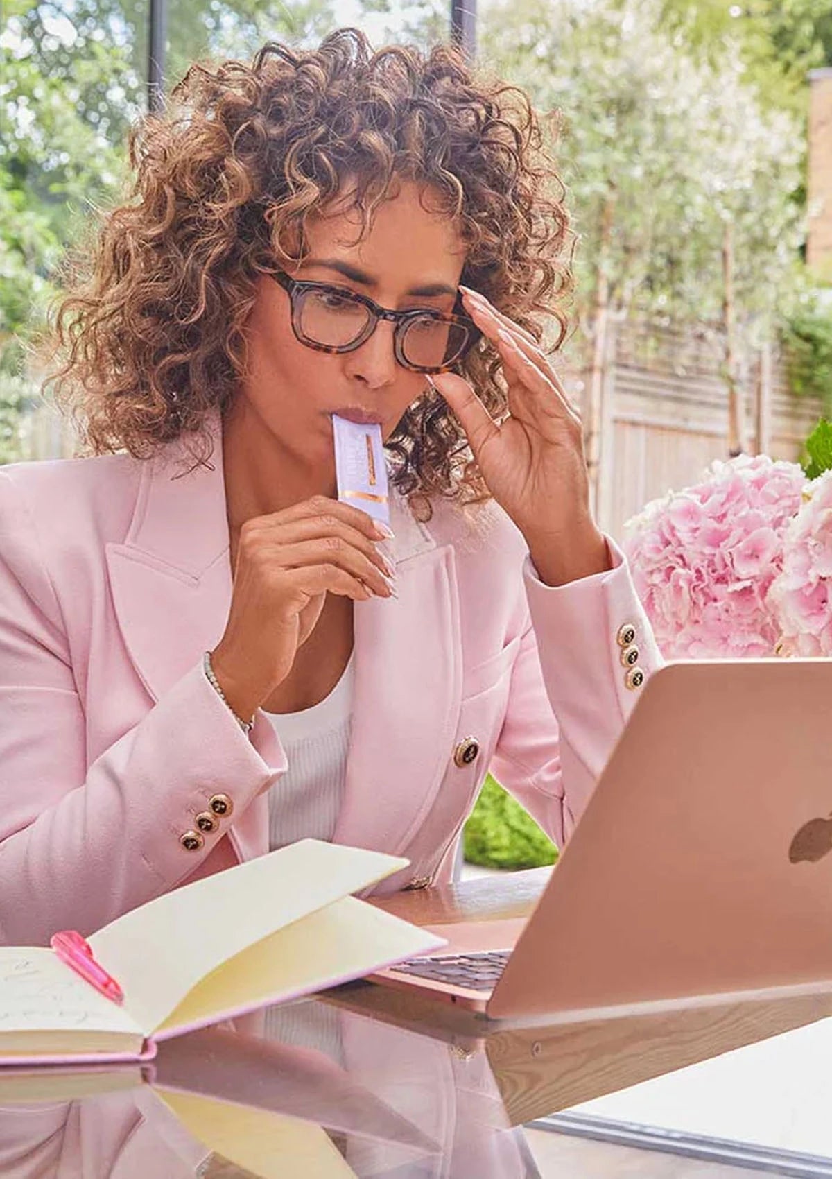 Woman in pink blazer sitting at a table with a laptop and notebook, outdoors.