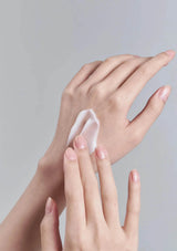 Two hands applying a white cream to another hand against a gray background