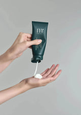 Hand holding a green tube labeled 'm' and squeezing white cream onto a palm against a gray background