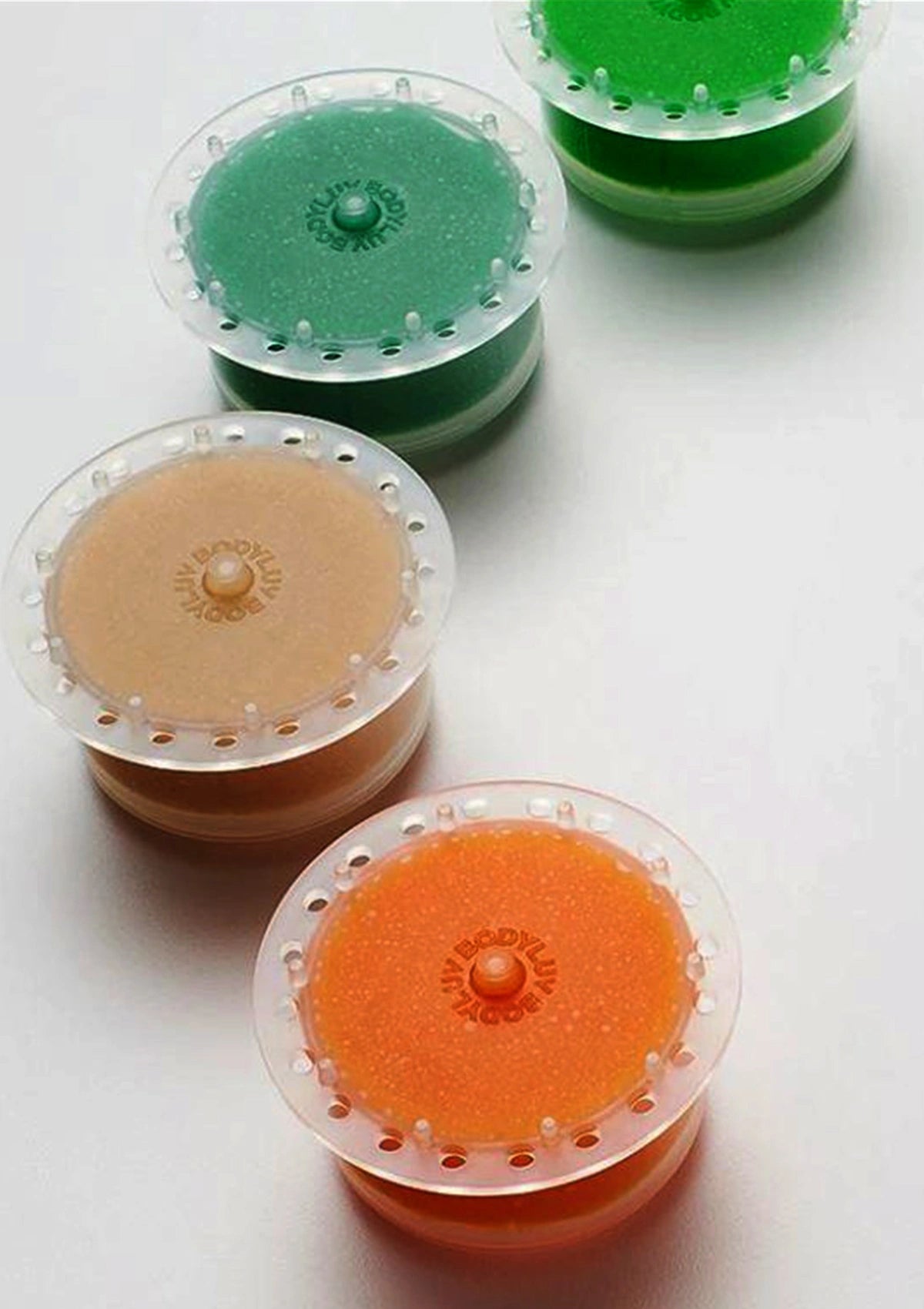Three small round gel plugs in translucent clear plastic with perforated edges, shown in green, peach-beige, and orange on a white background.