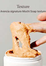 Mochi soap being held with a finger, showing its texture, against a gray background.