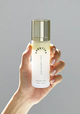 Hand holding a bottle of 'Prestige Collagen Age' cream mist against a gray background