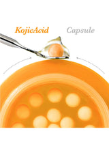Kojic Acid capsule being scooped with a spoon into a bowl of orange liquid.