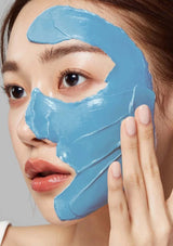 Woman applying blue facial mask to her face against a neutral background