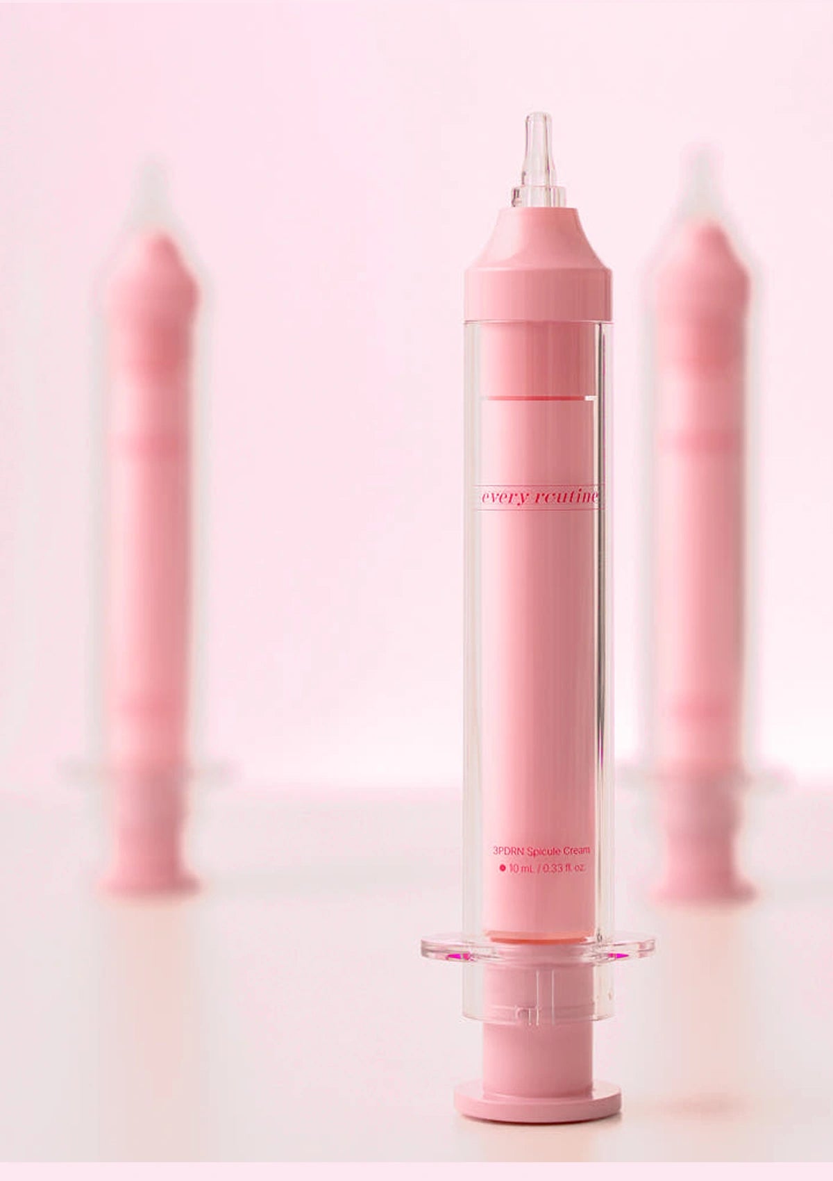 A pink and clear syringe-style cosmetic applicator standing upright on a stand, with three matching blurred applicators in the background against a soft pink studio backdrop.