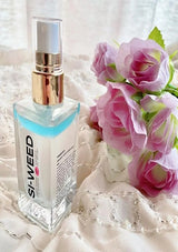 Clear bottle with gold cap labeled 'Si-Weed' next to pink flowers on a textured surface