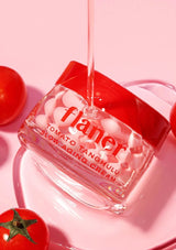 Flaner Tomato Tanghulu Slow Aging Cream being poured into a glass container with tomatoes on a pink background.