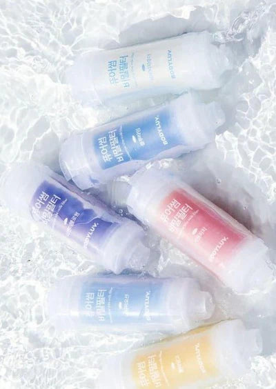 Six small cylindrical cosmetic tubes with translucent caps and white bodies, arranged on a textured white surface. The tubes come in white, blue, purple, pink, and pale yellow shades, with Korean text printed on the front.