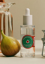 Clear glass bottle with dropper labeled 'Juliusy' next to a fig on a neutral background