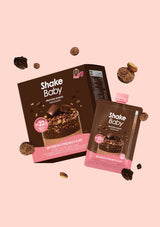 Shake Baby protein powder chocolate pouch and box packaging floating on a solid pale pink background, with scattered chocolate pieces around them. The packaging is dark brown with pink accents and white Shake Baby branding.